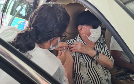 O apresentador Raul Gil levanta a manga de sua camisa para receber dose de vacina de enfermeira sentado em seu carro