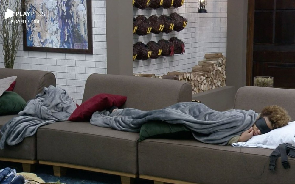 A peoa Lidi Lisboa dorme com um cobertor cinza e máscara de dormir na sala de A Fazenda 12