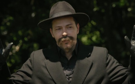 O ator Heitor Martinez com chapéu, luvas e sobretudo pretos caracterizado como Bernardo em cena de Amor Sem Igual