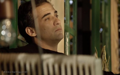 Sentado em um banco na varanda de casa, Rafael Sardão está com expressão triste como Miguel em cena de Amor Sem Igual