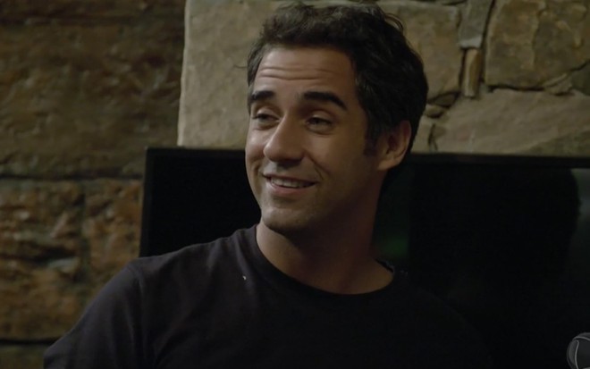 O ator Rafael Sardão em frente a uma lareira sorri como Miguel em cena de Amor Sem Igual