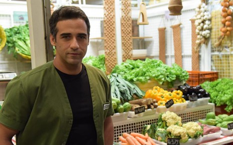 O ator Rafael Sardão caracterizado como o Miguel de Amor Sem Igual, com um colete verde e blusa preta, ele está em uma barraca de feira com várias frutas e hortaliças