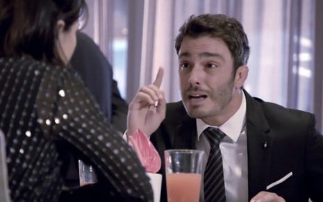 O ator Thiago Rodrigues com o dedo em riste diante de uma mesa com bebidas como Tobias em cena de Amor Sem Igual