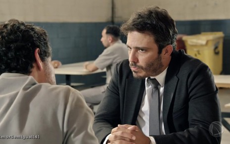 O ator Thiago Rodrigues caracterizado como Tobias com expressão de contrariedade sentado à mesa em uma cadeia em cena de Amor Sem Igual