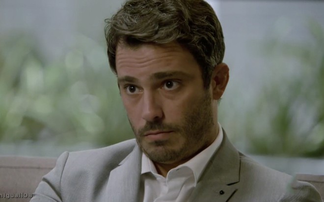 O ator Thiago Rodrigues sentado em um sofá como Tobias com uma expressão de surpresa em cena de Amor Sem Igual