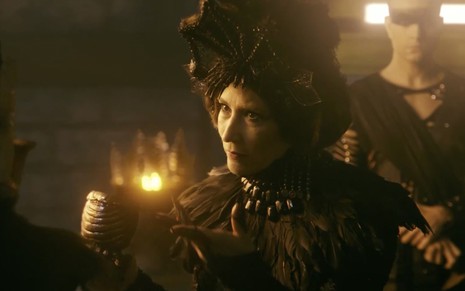 A atriz Beth Goulart como Jaluzi, com um ornamento na cabeça de um tamanho exagerado, feito de metal, enquanto segura um copo de metal com a mão direita em cena de Gênesis
