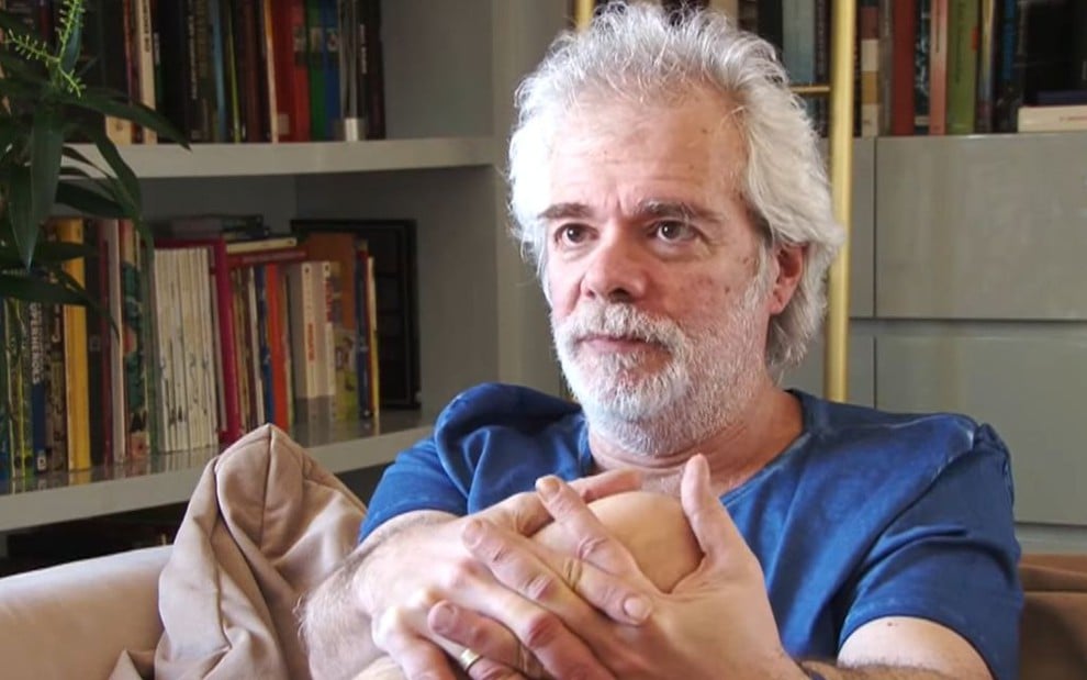 O autor Carlos Lombardi, na sala de sua casa, segura o joelho com as duas mãos