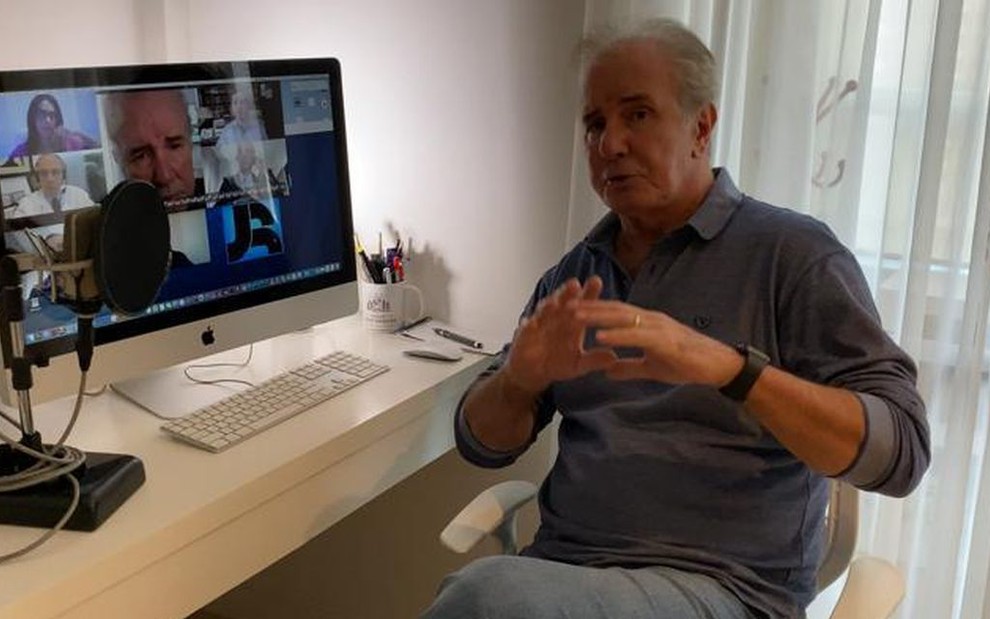 Com camisa social azul, Celso Freitas está sentado diante de um computador e de um microfone profissional