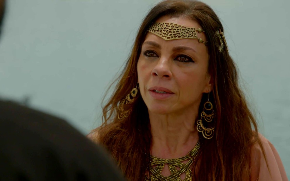 Carla Marins grava cena de Gênesis com acessório dourado na cabeça, na orelha e no pescoço e expressão de tristeza como Adália