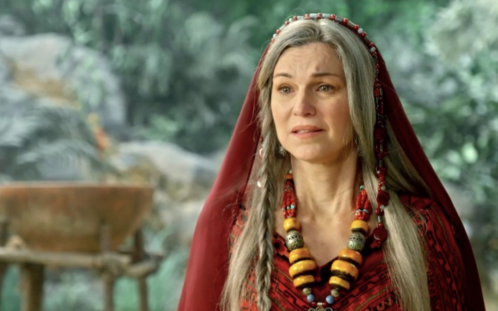 A atriz Adriana Garambone como Sarai, com uma roupa vermelha ricamente adornada por joias, com expressão de confusão em cena de Gênesis