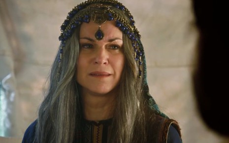 A atriz Adriana Garambone como Sarai com cabelos brancos, aparência envelhecida e uma roupa de festa, ricamente adornada com pedras azuis, em cena de Gênesis