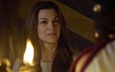 A atriz Adriana Garambone como Sarai com os cabelos soltos está dentro de uma tenda como Sarai, há a chama de uma lampião em primeiro plano e uma mulher de costas, à direita, em cena de Gênesis