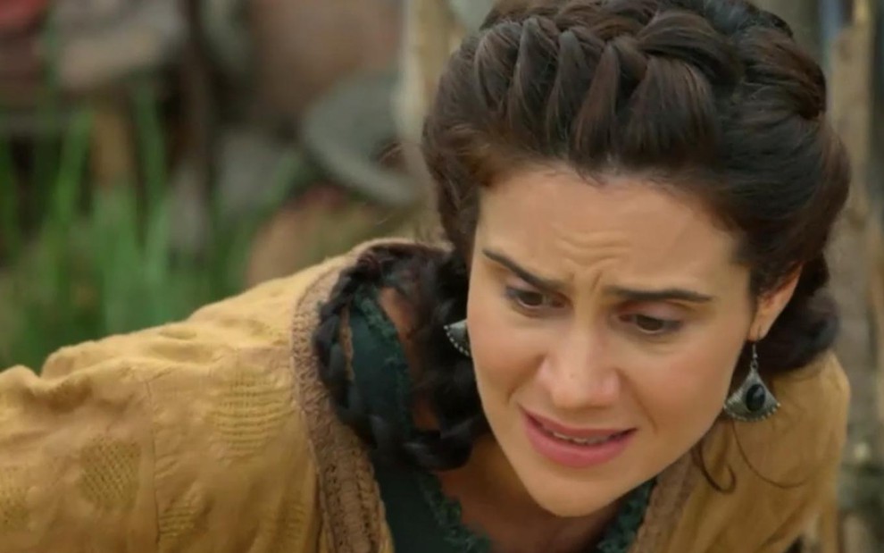 Branca Messina em cena de Gênesis: caracterizada com Amat, atriz olha com desespero para o chão e faz expressão de dor