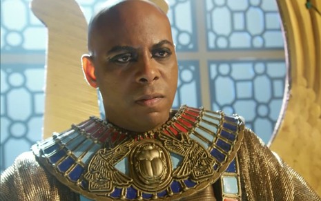 O ator André Ramiro careca e com roupas do Egito Antigo, com um olho de Hórus no peito e joias vermelhas e turquesas pelo corpo, em cena de Gênesis