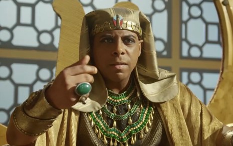 O ator André Ramiro sentado em um trono com roupas do Egito Antigo, coberto de joias e ouro, como o faraó Amenemhat 3º em cena de Gênesis