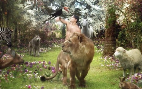 O ator Carlo Porto está nu em um cenário virtual do jardim do éden, com vários animas como um leão, um tucano, um cavalo branco ao seu redor em cena de Gênesis