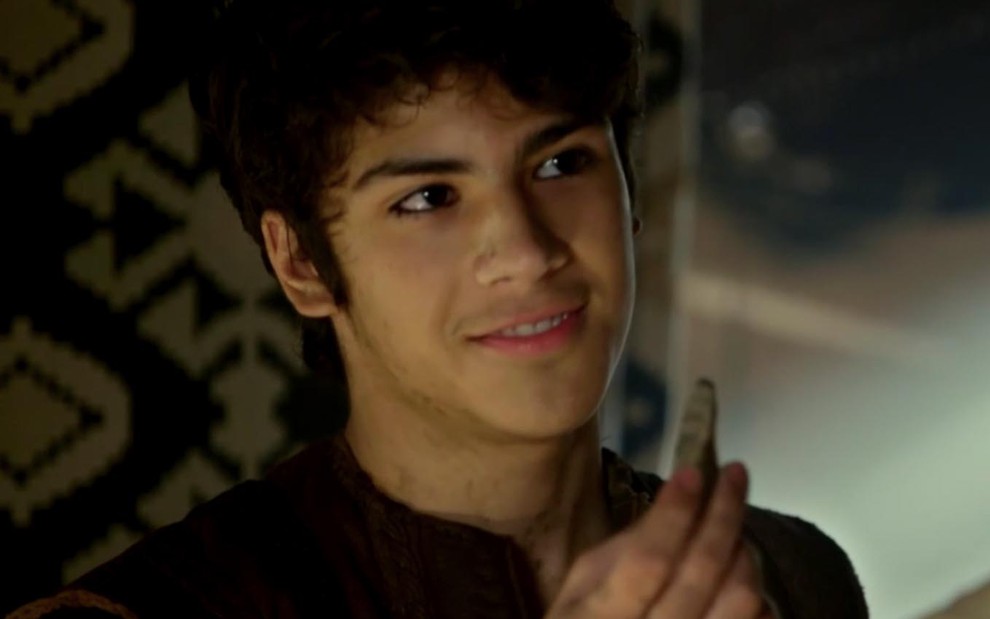 O ator Henrique Camargo como Ismael, com expressão maliciosa, está segurando uma peça de bronze com a ponta dos dedos da mão direita em cena de Gênesis