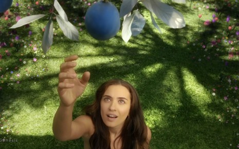 A atriz Julliana Boller nua como Eva colhe um fruto azul do tamanho de uma goiaba em cena de Gênesis