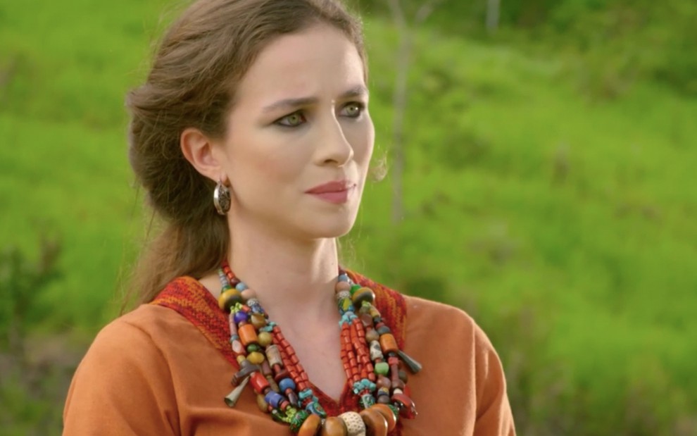 A atriz Laryssa Alves caracterizada como Sarai, com uma túnica laranja, colares de contas coloridas e brincos de argola dourados, na fase Ur dos Caldeus em Gênesis