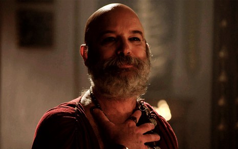 Jairo Mattos em cena de Gênesis: caracterizado como Morabi, ator coloca a mão no peito e sorri com orgulho para alguém fora do quadro