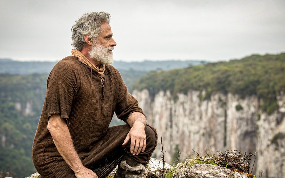 O ator Oscar Magrini ajoelhado a beira de um cânion caracterizado como Noé em cena de Gênesis