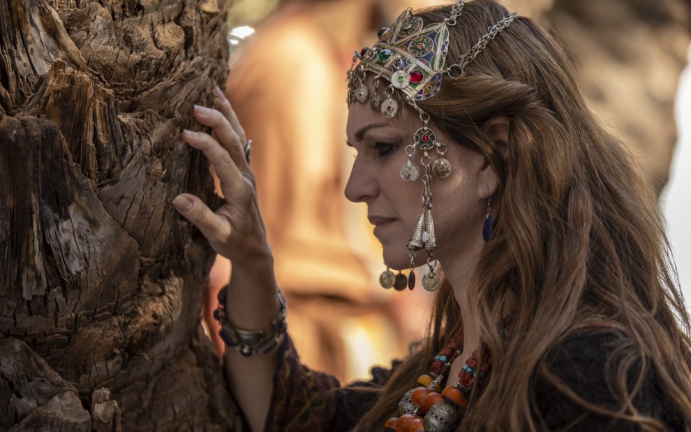 Adriana Garambone grava com acessório na cabeça e mãos apoiadas em uma árvore como Sarai de Gênesis