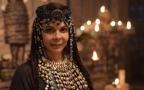 A cantora Sula Miranda como Quirá, com um adorno de contas na cabeça e um colar com pérolas que desce pelo tronco, dentro de um palácio em cena de Gênesis