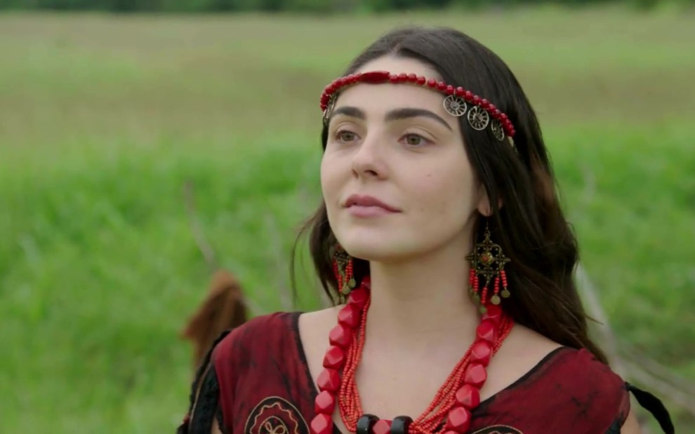 Marcela Barrozo grava com colar e acessório na cabeça vermelhos e vestido vermelho e preto como Uriala de Gênesis