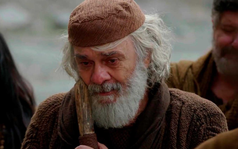 O ator Zécarlos Machado segura uma bengala de madeira junto ao corpo, com longos cabelos e barba brancos, caracterizado como Abraão em cena de Gênesis