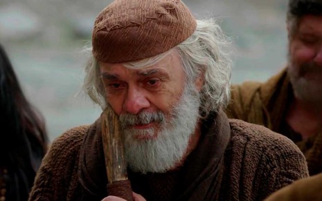 O ator Zécarlos Machado segura uma bengala de madeira junto ao corpo, com longos cabelos e barba brancos, caracterizado como Abraão em cena de Gênesis
