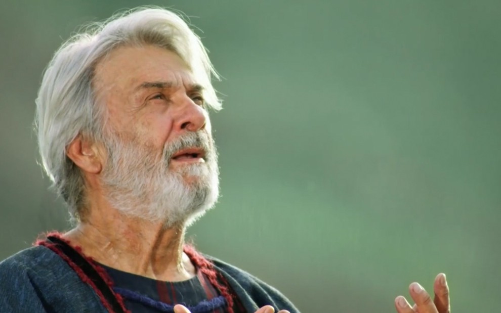 O ator Zécarlos Machado está em um campo verdejante e olha para cima, como se orasse, no papel de Abrão em cena de Gênesis