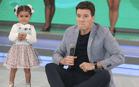 Uma criança de cinco anos está em pé à esquerda, enquanto Rodrigo Faro está sentado no palco do Hora do Faro com expressão de contragosto