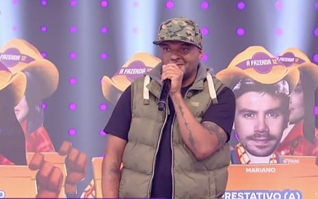 O rapper e beatboxer Fernandinho Beat Box no palco do Hora do Faro com um microfone nas mãos e caricaturas dos peões de A Fazenda ao fundo