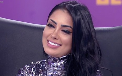 MC Mirella sentada em uma poltrona preta com um sorriso sem graça no palco do Hora do Faro