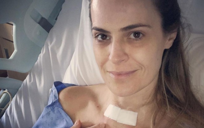 Regiane Tápias em cama de hospital aponta para marca de sua cicatriz após cirurgia coberta com curativo