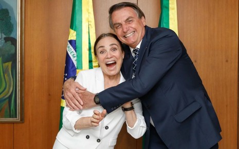 Regina Duarte de boca aberta sendo abraçada pelo presidente Jair Bolsonaro