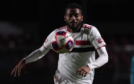 Reinaldo com uniforme branco do São Paulo, com faixas vermelha e preta na altura do peito, olha para a bola quase na altura de seu rosto