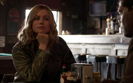 A atriz Brie Larson faz cara de pensativa com o balcão apoiado em uma mesa de bar, com Samuel L. Jackson de costas, em cena de Capitã Marvel