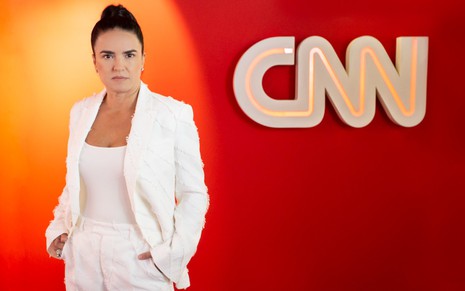 Renata Afonso com uma blusa, terno e calça brancos, com as mãos no bolso, em uma parede vermelha com o logotipo da CNN