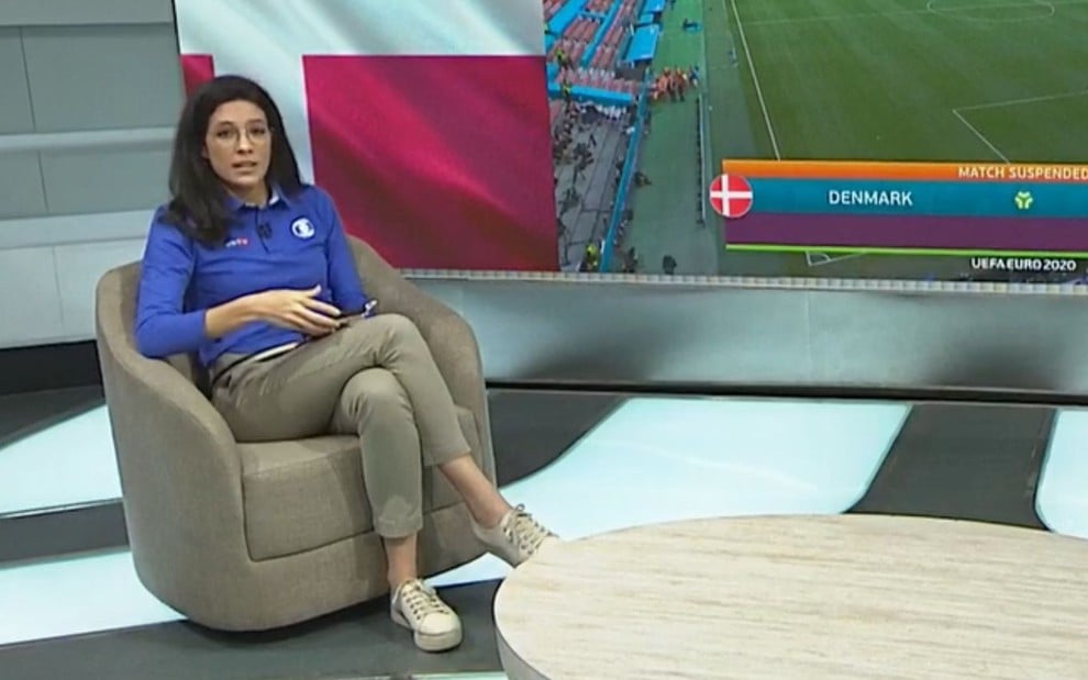 Em uma poltrona caqui, a narradora Renata Silveira está sentada de pernas cruzadas em frente a um telão com imagens do jogo ao fundo