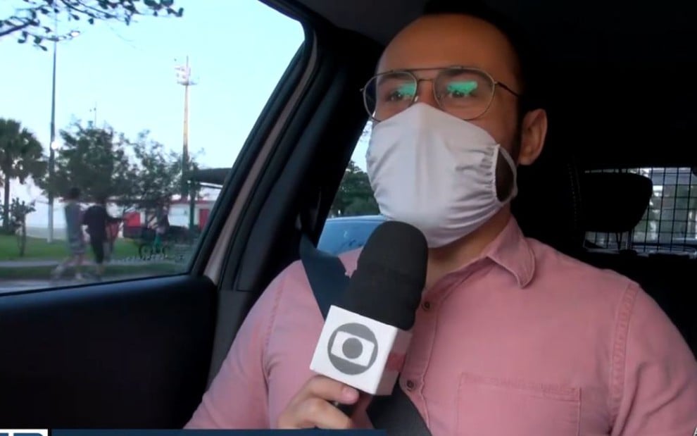 Rômulo D'Avila dentro de um carro, de camisa social rosa, óculos de grau e máscara branca