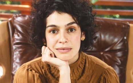A atriz Maria Flor, com a mão no queixo, sentada em uma poltrona