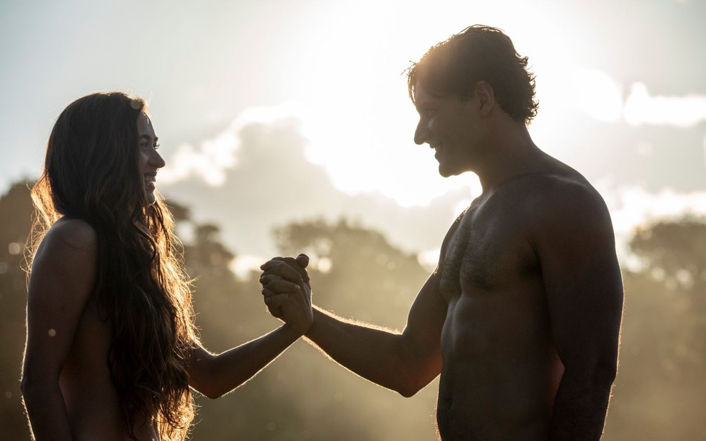 Os atores Juliana Boller e Carlo Porto dão as mãos, sem roupas, em cena ensolarada de Gênesis