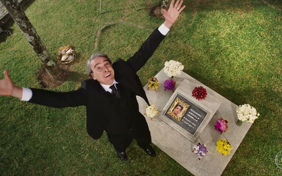 Aparício (Alexandre Borges) com os braços para cima comemorando diante de um túmulo em cena de Haja Coração