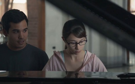 Guto (Bruno Gadiol) sentado ao lado de Benê (Daphne Bozaski) no piano em cena de Malhação