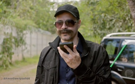 Com bigode falso, óculos escuros e boné, Heitor Martinez fala ao celular em cena de Amor Sem Igual