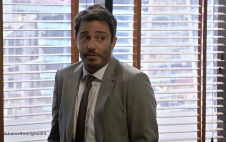 O ator Thiago Rodrigues de terno e gravata em cenário de escritório na novela Amor Sem Igual