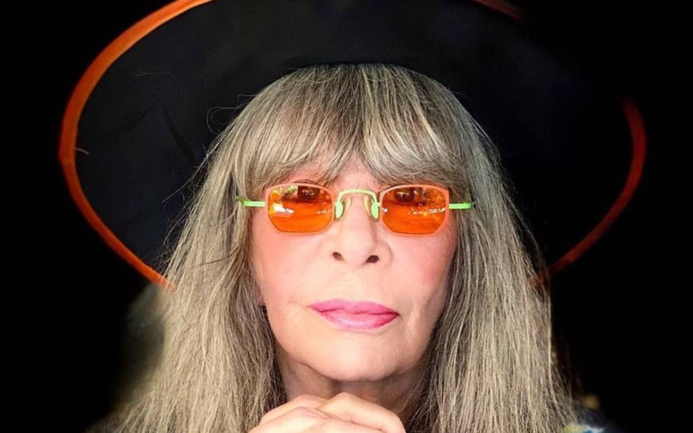 Rita olha para frente, usa óculos laranja, chapéu preto e está com o cabelo solto
