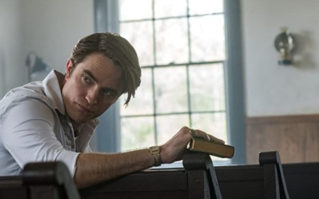 Robert Pattinson sentado em igreja e segurando uma Bíblia em cena de O Diabo de Cada Dia (2020), filme da Netflix