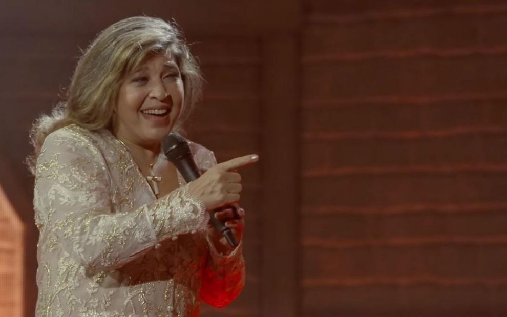 Roberta Miranda com roupa dourada e cabelo solto segurando microfone e apontando para frente